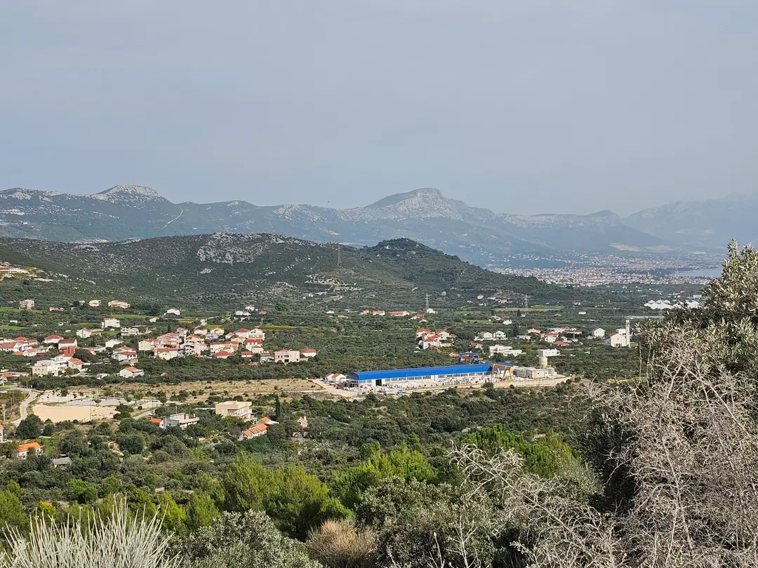 Pogon u Planu snimljen s brda: hala s dizalicom, selo oko nje, kršni greben i Kaštela s morem na obzoru