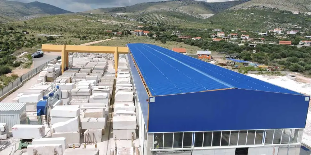 The Sprega Kamen plant from the air: the blue-roofed hall, the yellow gantry crane and a yard full of sawn blocks, with the village of Plano and the ridge behind.